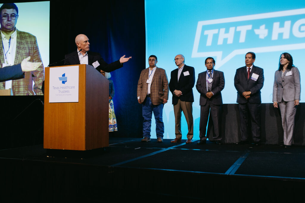 Speaker at a wooden podium on a stage microphone in front with a large blue backdrop and several suited individuals standing to the right of the podium at a conference The podium badge reads Texas Healthcare Trustees