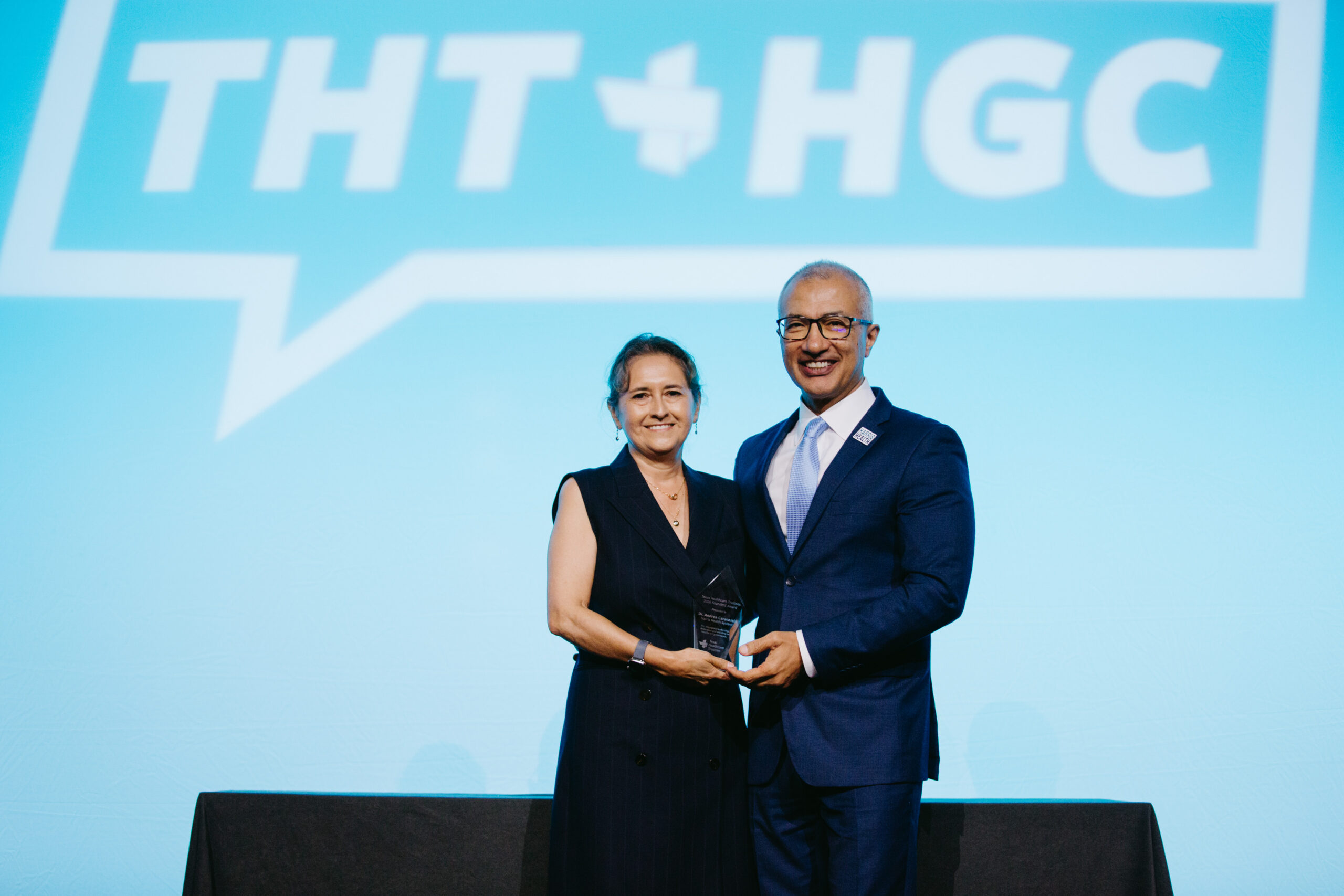 06052026_HGC_Awards Luncheon_nguillot-21 - Texas Healthcare Trustees Two professionals in dark suits pose on stage holding a glass award against a blue backdrop with large white lettering behind them