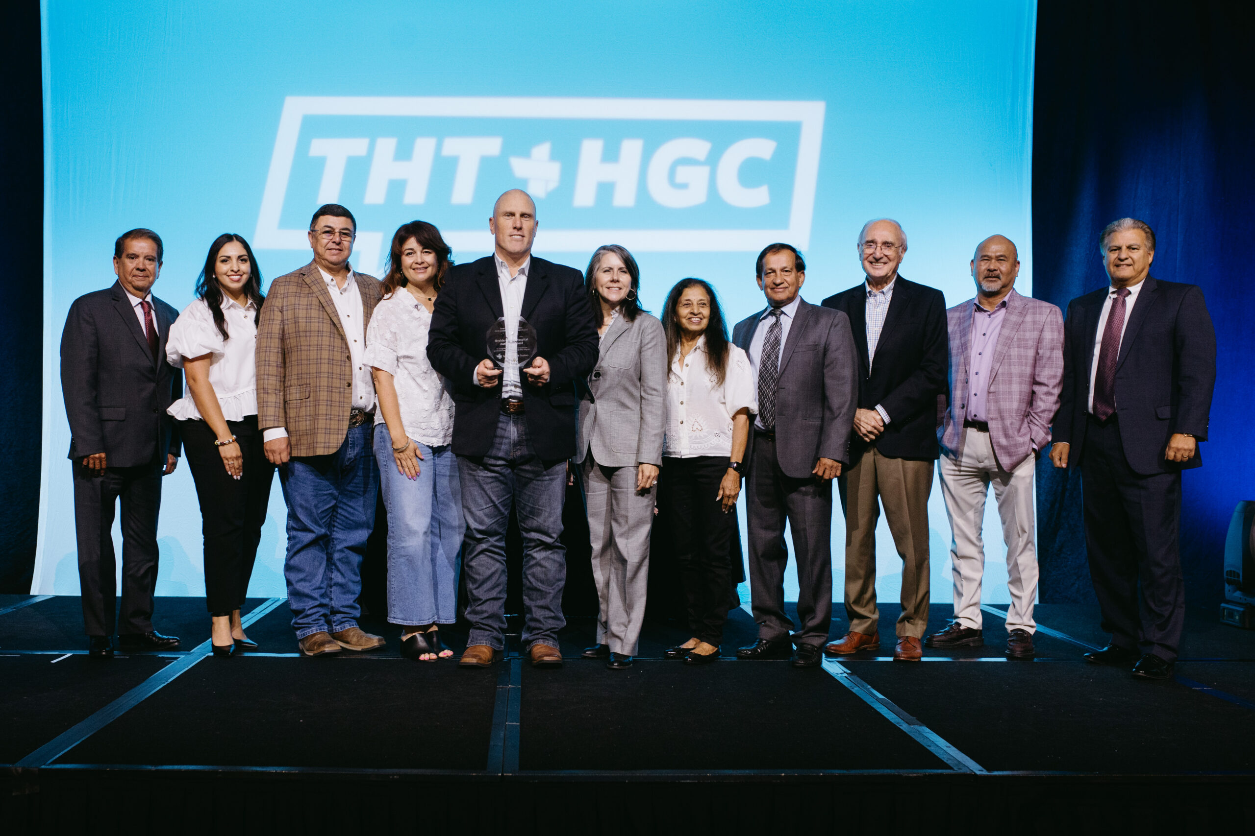Group of eleven adults on a stage posing for a photo the man in the center holds a glass trophy while others smile with a blue backdrop and logo behind them