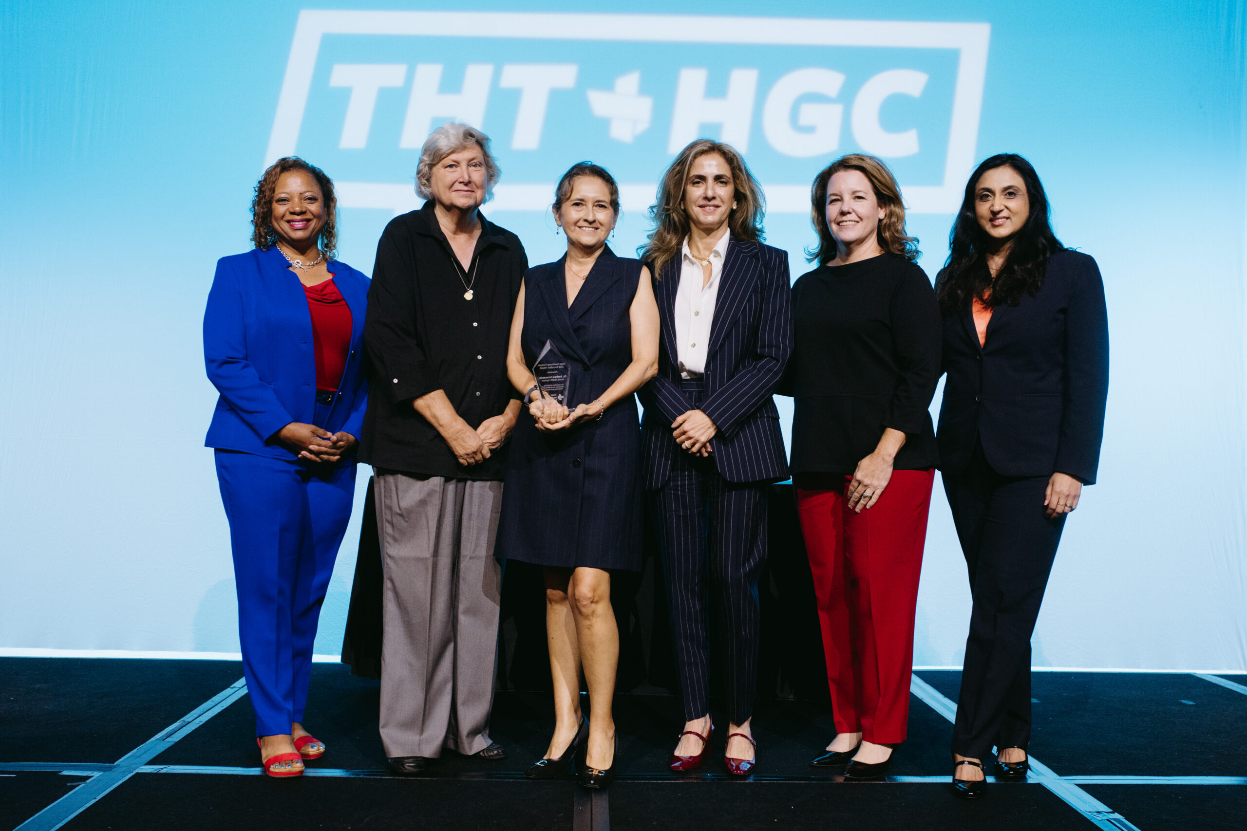 06052026_HGC_Awards Luncheon_nguillot-23 - Texas Healthcare Trustees Six women in formal business wear pose together on a stage with one holding a glass award at center against a blue backdrop with a logo