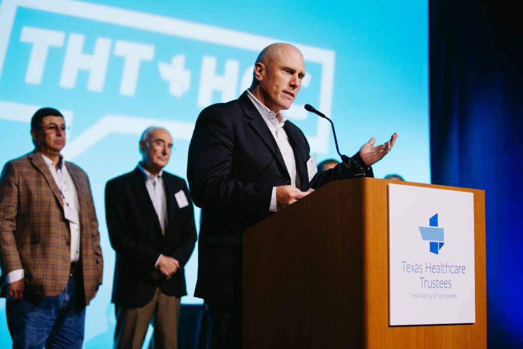 Speaker at a podium delivering a talk with three suited colleagues in the background on a stage blue backdrop featuring a large logo The podium sign reads Texas Healthcare Trustees