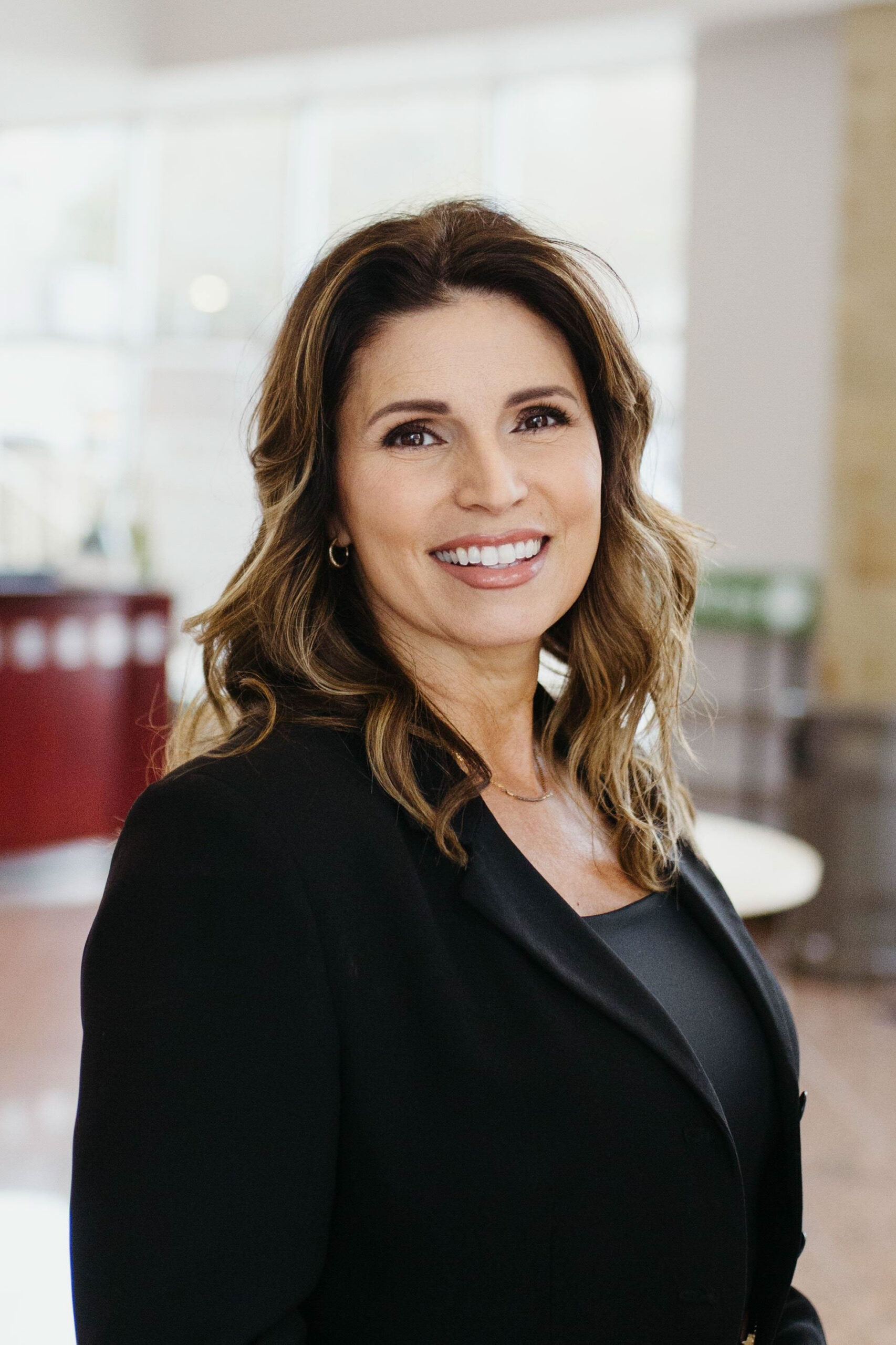 Smiling woman with shoulder length wavy brown hair wearing a black blazer in a bright indoor setting
