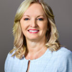Headshot_Carden_Carol - Texas Healthcare Trustees Smiling woman with blonde hair wearing a light blue textured jacket posing against a gray backdrop head and shoulders