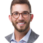 Headshot_Mckinney_Dillon - Texas Healthcare Trustees Professional headshot of a smiling man with brown hair and a beard wearing glasses and a gray blazer