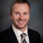 Headshot_Wysocki_Keith - Texas Healthcare Trustees Smiling man in a dark blazer white shirt and plaid tie head and shoulders portrait against a gray gradient background