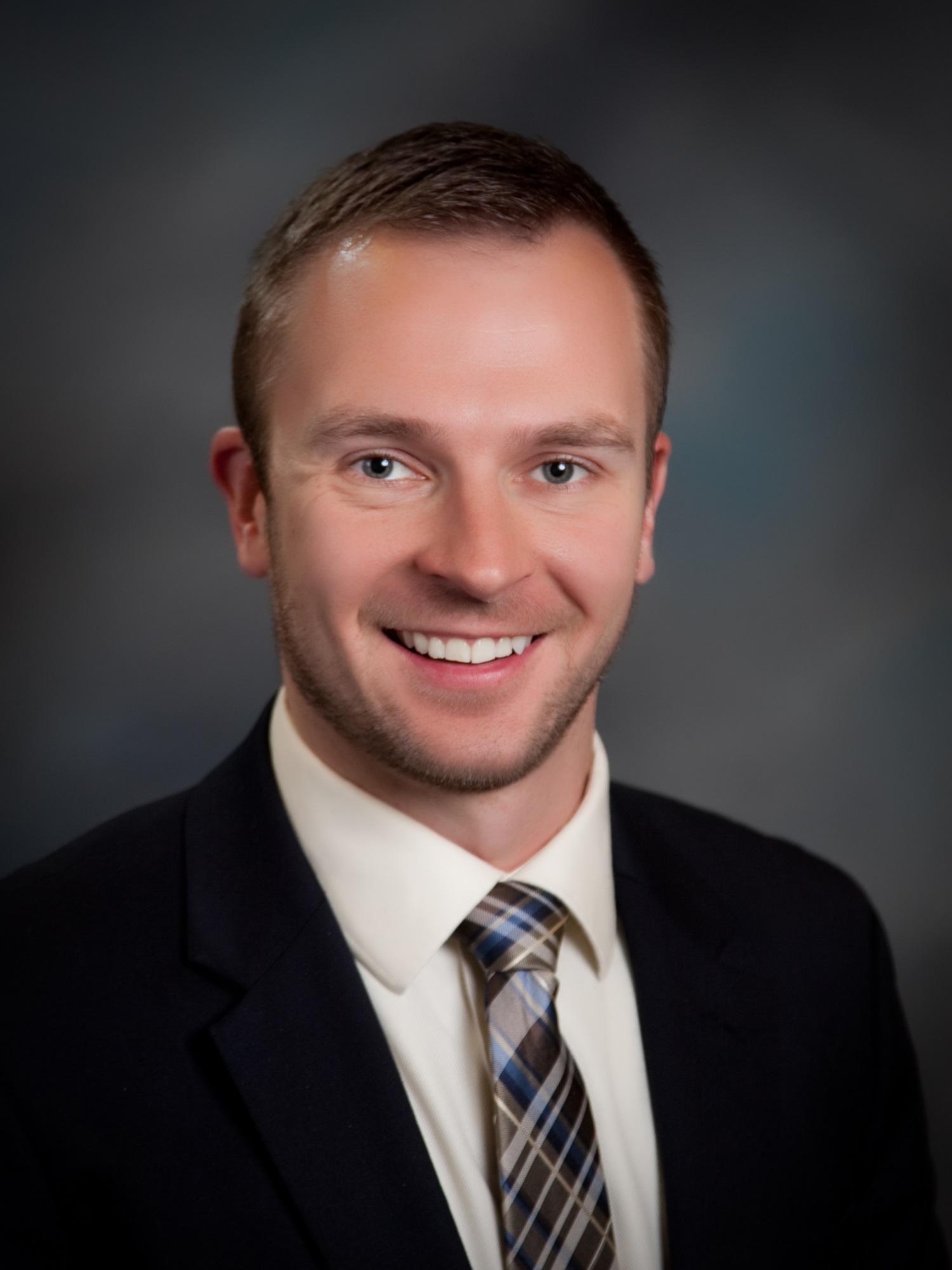 Smiling man in a dark blazer white shirt and plaid tie head and shoulders portrait against a gray gradient background