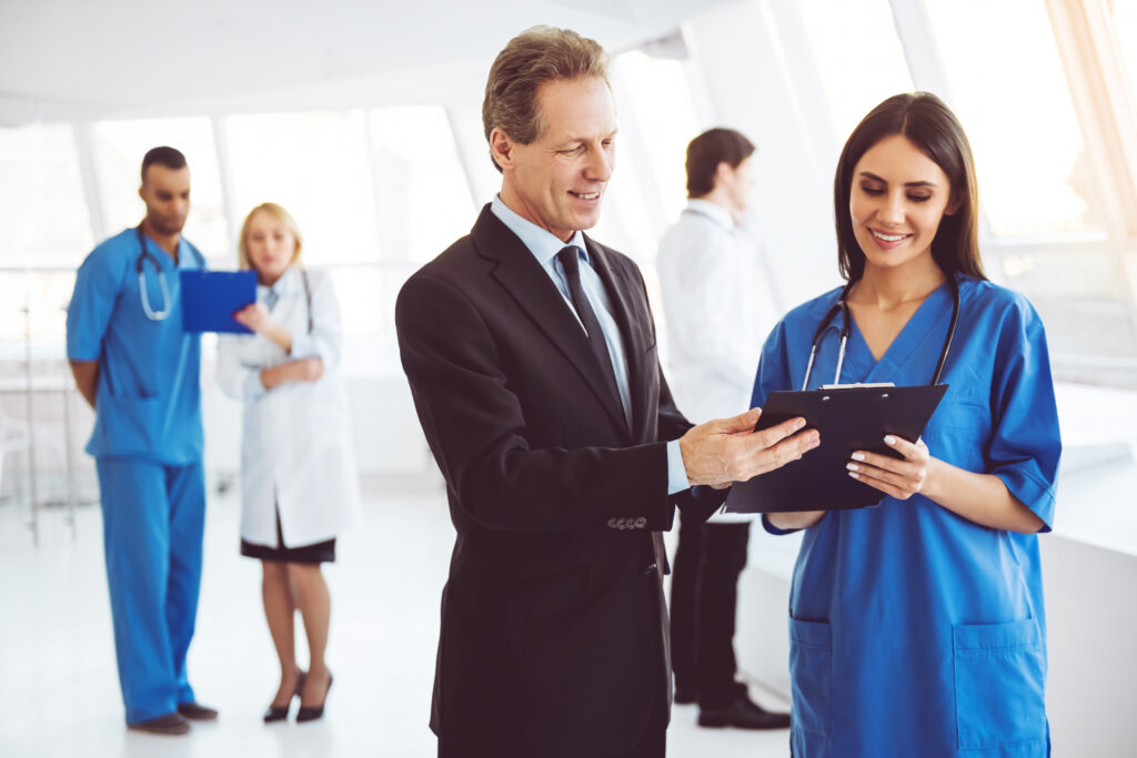 Doctor and businessman - Texas Healthcare Trustees A man in a suit shakes hands with a smiling nurse in blue scrubs holding a clipboard in a bright hospital lobby