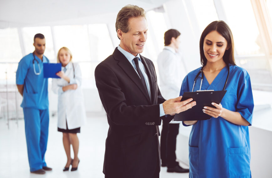 A man in a suit shakes hands with a smiling nurse in blue scrubs holding a clipboard in a bright hospital lobby.