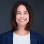 Jamie Singer - Texas Healthcare Trustees Head and shoulders portrait of a smiling woman with shoulder length dark hair wearing a navy blazer and white blouse against a dark grey background