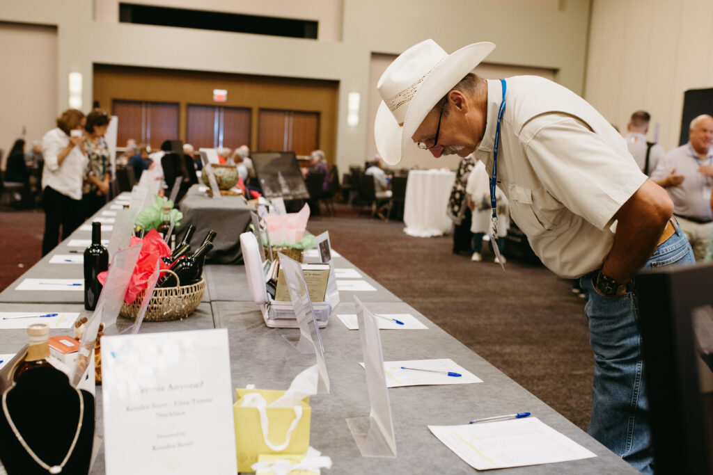 Man in a white cowboy hat and shirt leaning over a long table of display items at an indoor event hall inspecting goods as others browse in the background