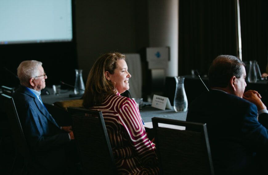 Smiling woman in a maroon striped dress at a conference table, with two men nearby in a dim meeting room.