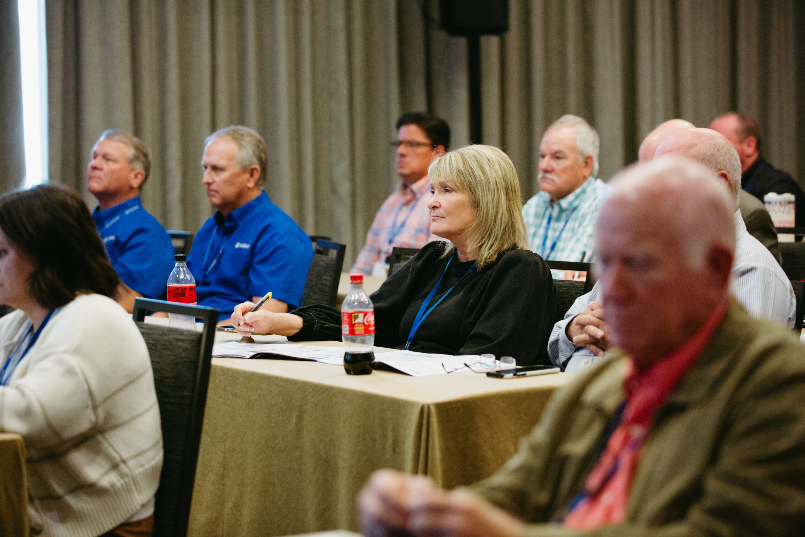 Group of adults in a conference room listening attentively; a blonde woman at the front table is taking notes with notebooks and beverages nearby.