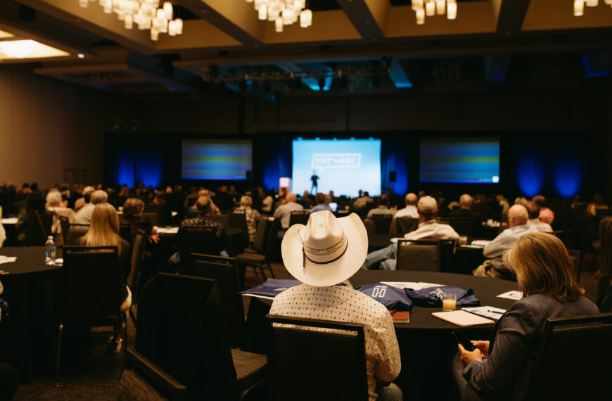 Conference hall full of attendees facing a stage with a speaker and a large logo screen behind them.