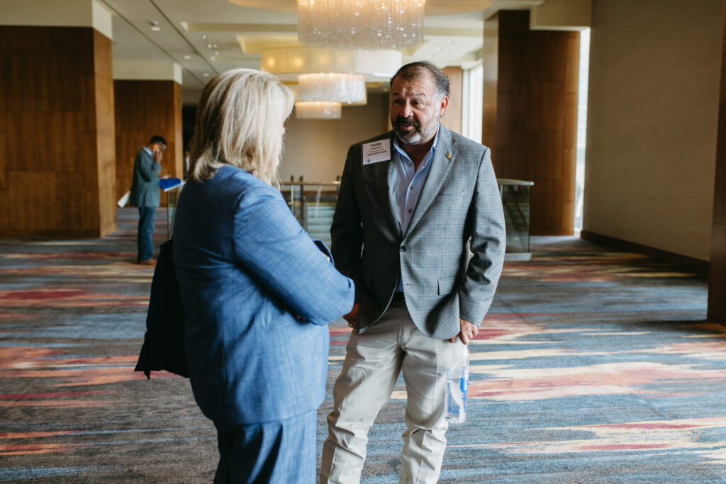 Man in a gray blazer converses with a woman in a blue jacket in a hotel lobby with chandeliers overhead