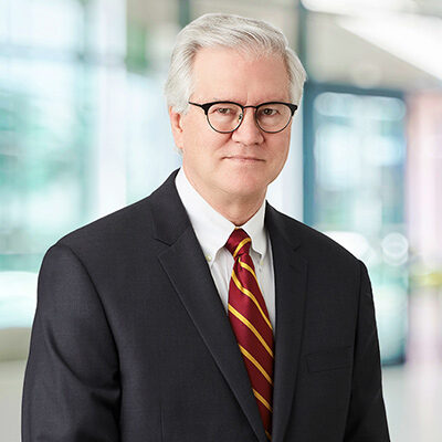 Professional portrait of an older man in a dark suit and striped red tie wearing glasses in a bright office setting