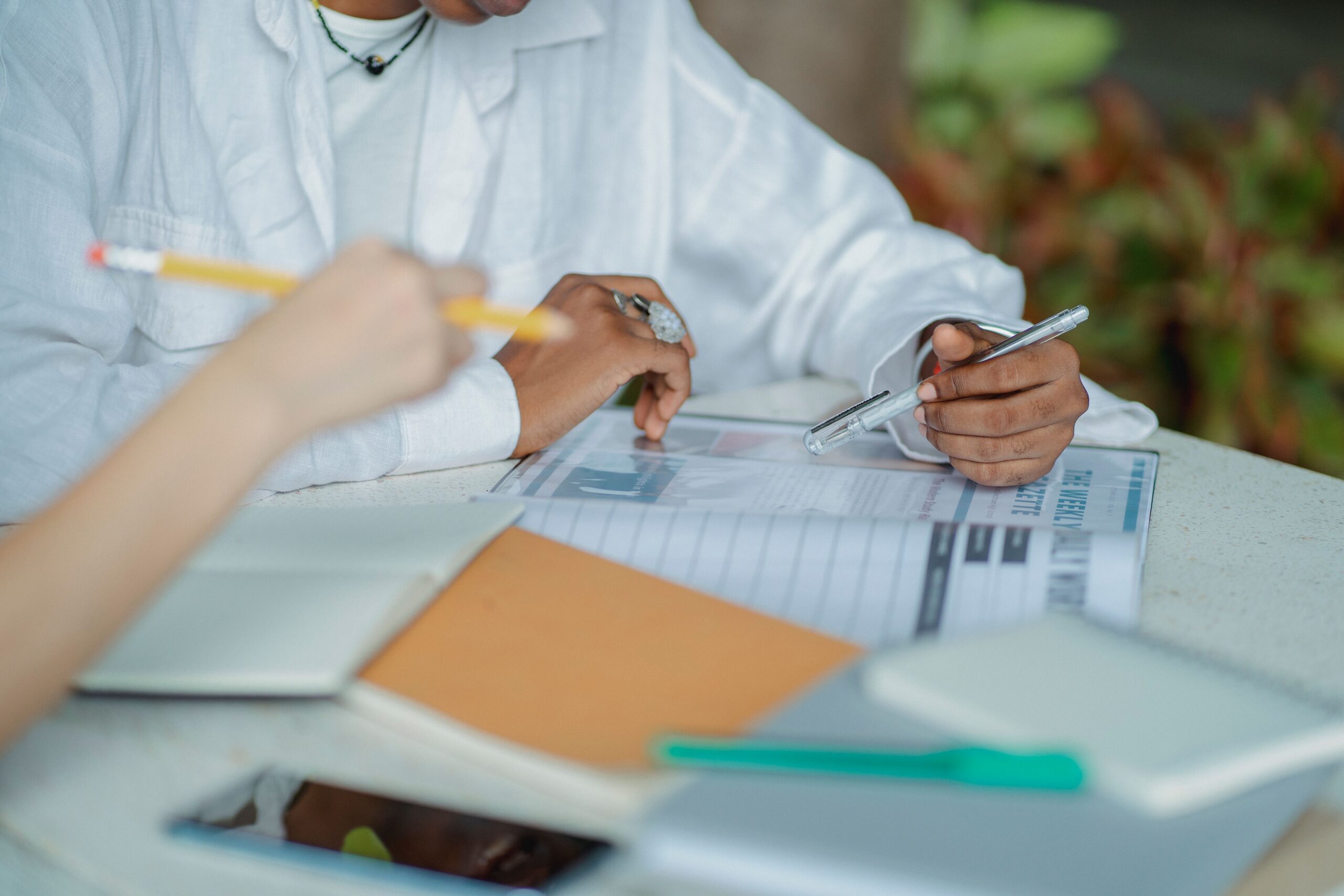 Two people sit at a table reviewing documents; one writes with a pencil while the other holds a phone.