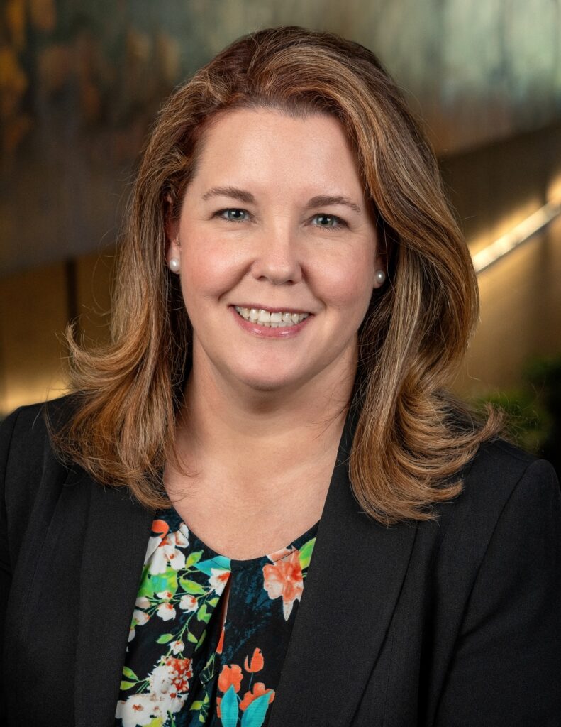 Carolynn Jones Official Headshot - Texas Healthcare Trustees Head and shoulders portrait of a smiling woman with light brown hair wearing a black blazer and a colorful floral blouse with pearl earrings on a blurred backdrop